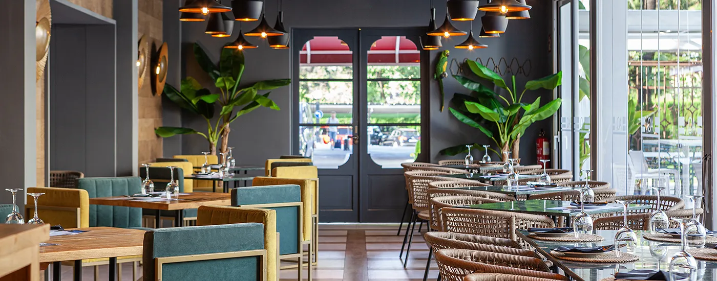 Comedor moderno con mesas, sillas de colores y barandilla con plantas y l&aacute;mparas colgantes.