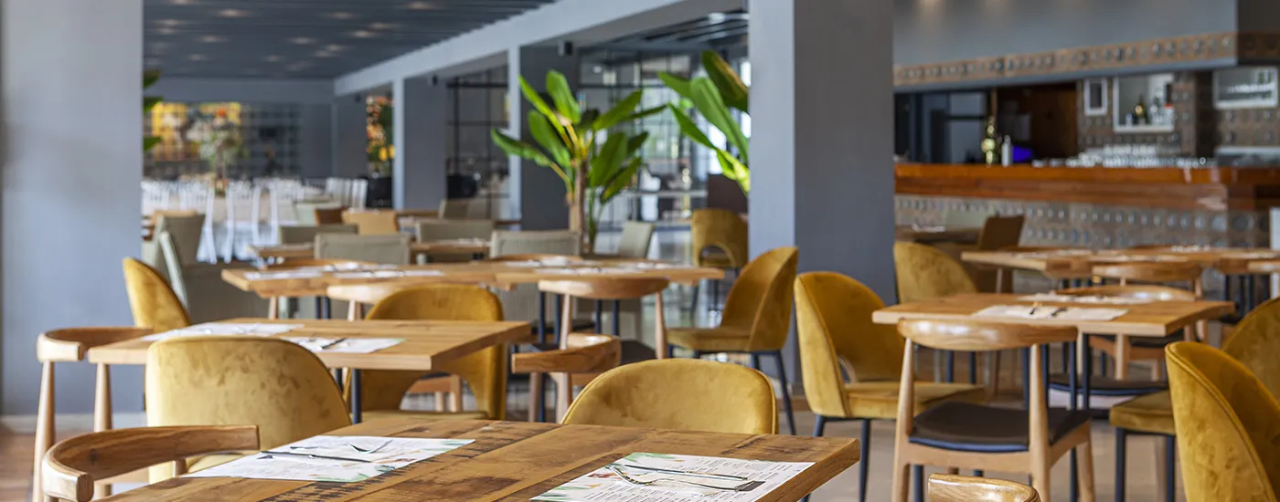 Interior de cafeter&iacute;a con mesas de madera y sillas doradas, planta al centro