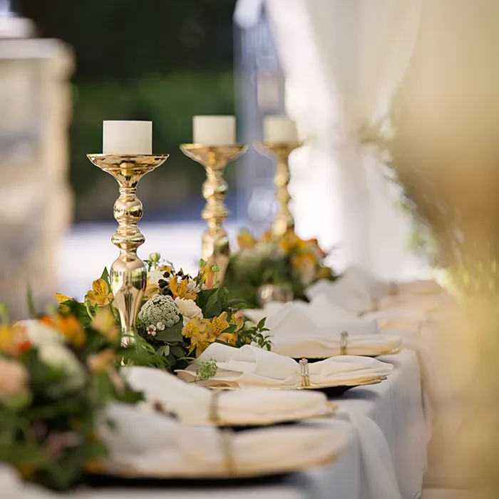 Mesa de banquete con velas, flores y cubiertos sobre mantel blanco.