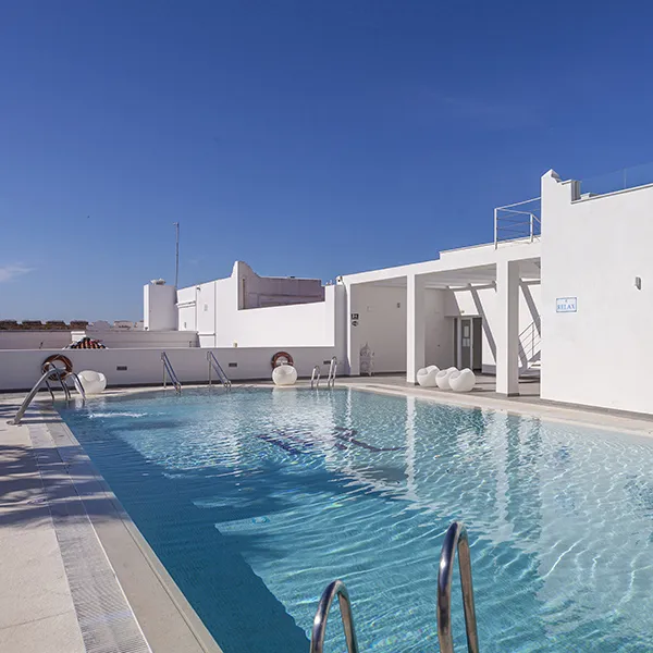 Piscina azul con edificio blanco y tumbonas, cielo despejado.