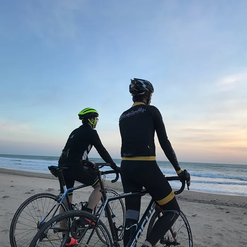 Dos ciclistas con casco en la playa junto al mar al atardecer.