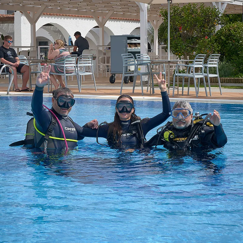Tres buceadores con aletas y trajes de neopreno saludan dentro de una piscina.