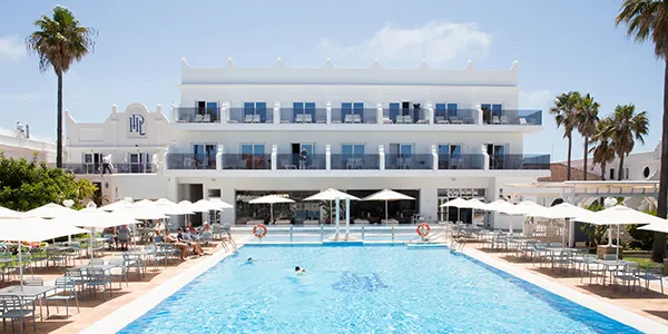 Hotel frente a piscina azul con tumbonas y sombrillas, palmeras al lado