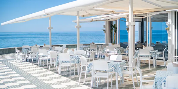 Terraza con mesas y sillas blancas frente al mar, bajo toldos.