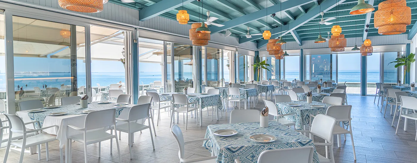 Interior de restaurante con mesas, sillas y luz colgante sobre el mar.