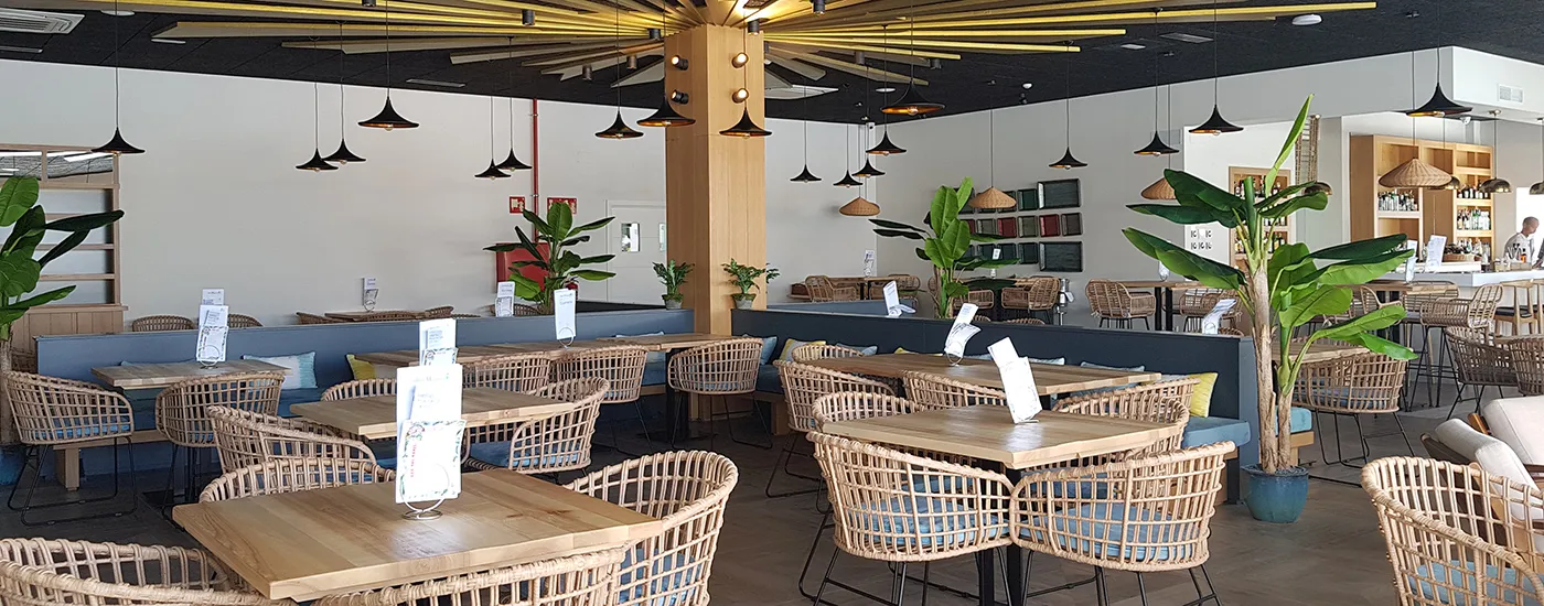Comedor moderno con mesas de madera, sillas de mimbre y plantas en interior.