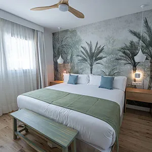 Dormitorio moderno con cama doble, palmeras y l&aacute;mpara de pie.