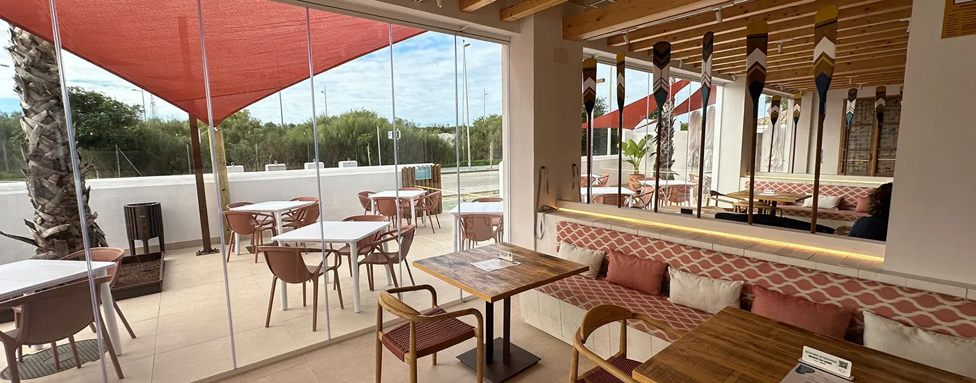 Terraza interior con mesas, sillas y ventanales, &aacute;rea de descanso con sillones