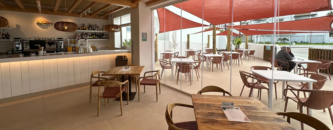 Cafeter&iacute;a con mesas, sillas y barra, luz natural y techo de madera.