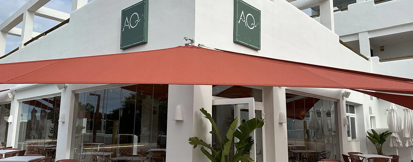 Entrada de edificio con toldo rojo, logo ACQUA y palmeras en terraza.