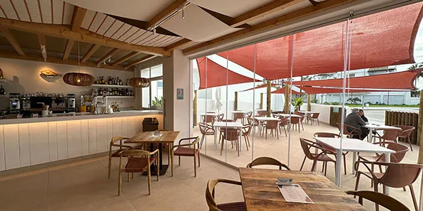 Cafeter&iacute;a con mesas y barra, terraza con toldos rojos y mesas al aire libre.