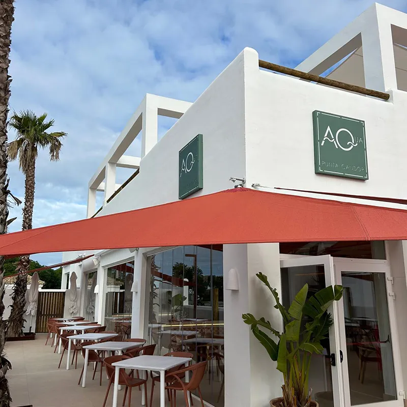 Terraza de bar con sillas, toldo rojo y fachada blanca con letreros verdes bajo cielo azul.