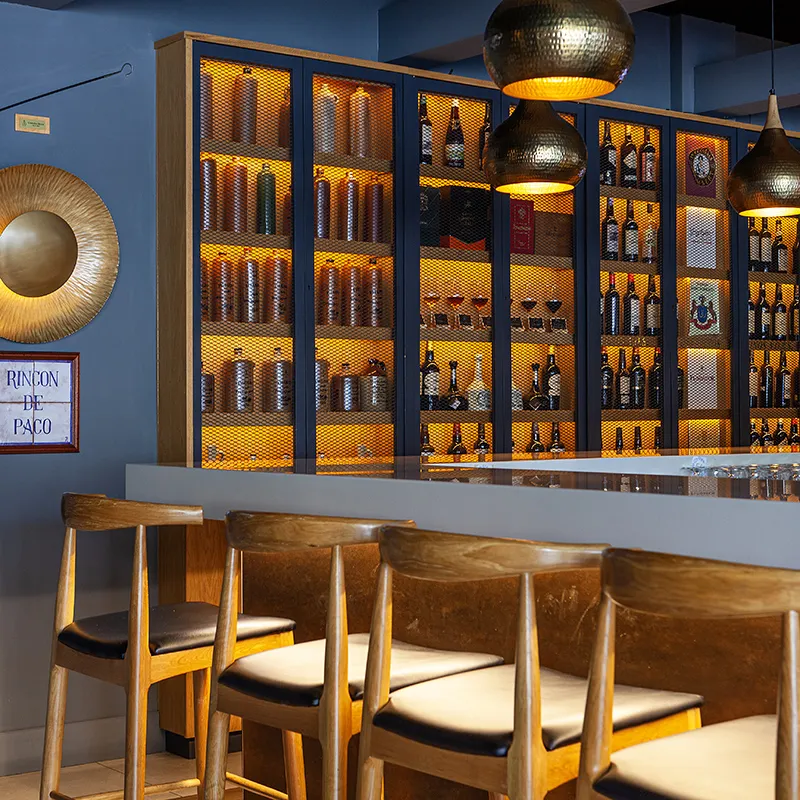 Bar con estanter&iacute;a de botellas, sillas de madera e iluminaci&oacute;n dorada.