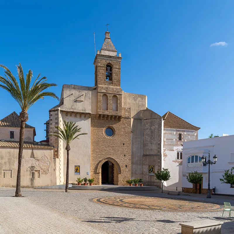 Iglesia de piedra con campanario y palmeras bajo cielo azul, plaza urbana