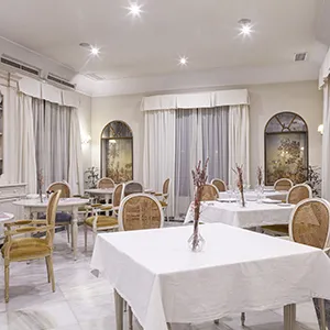 Comedor elegante con mesas blancas, sillas de rat&aacute;n y luz cenital.