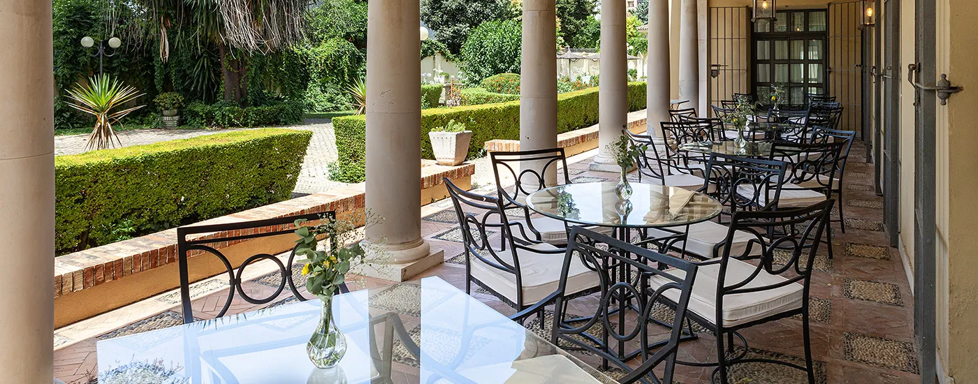 Terraza con mesas y sillas de metal, jard&iacute;n verde y columnas de piedra