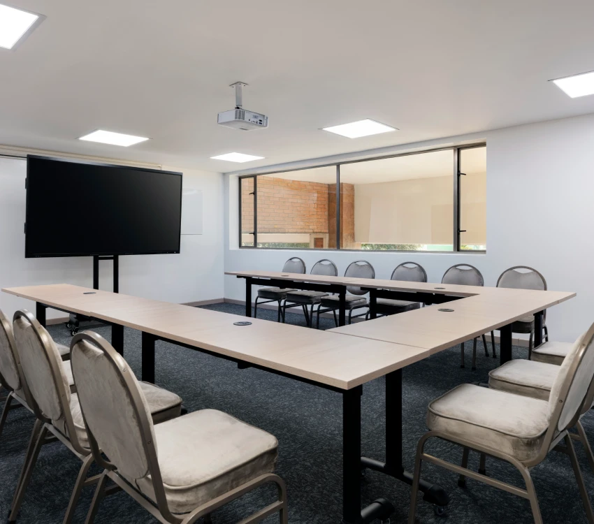 Sala de reuniones vac&iacute;a con mesa en forma de U, sillas y pantalla grande.