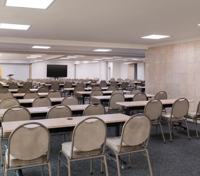 Sala de conferencias vac&iacute;a con mesas, sillas y pantalla grande al fondo.