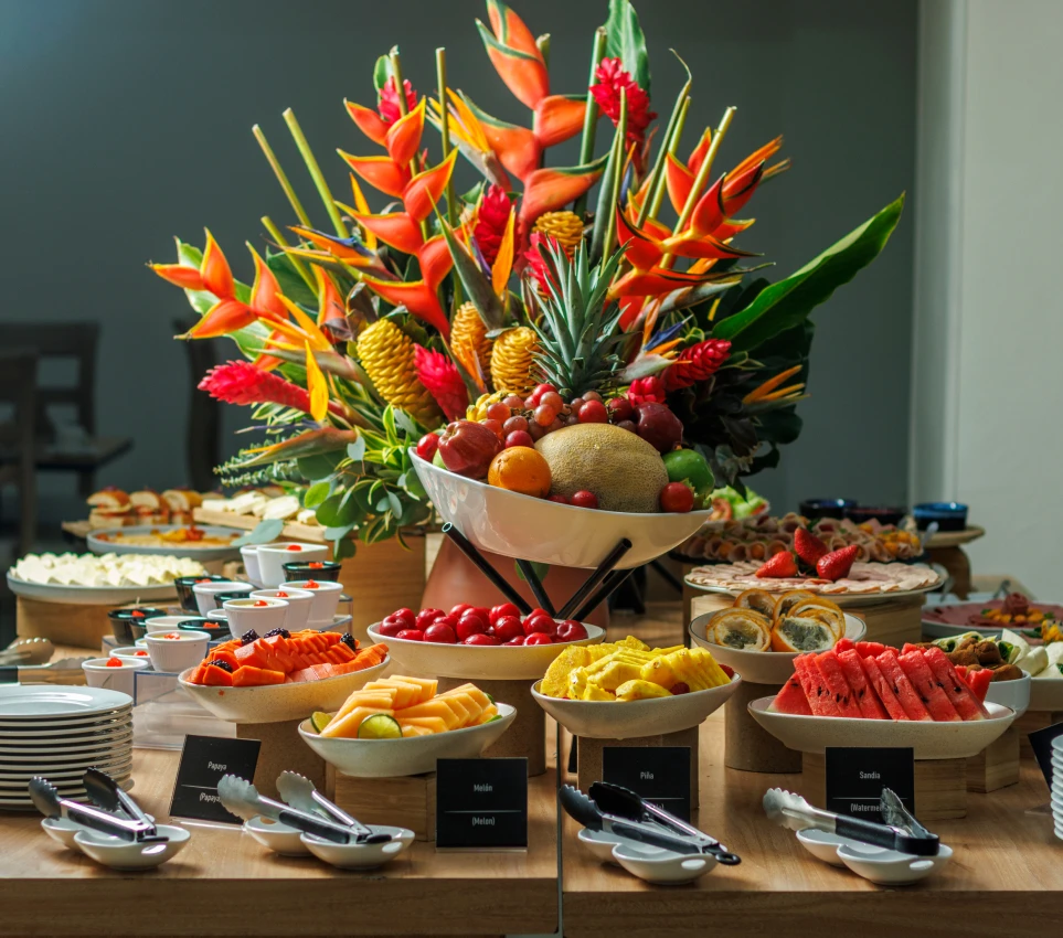 Mesa de buffet con flores tropicales y frutas variadas en platos.
