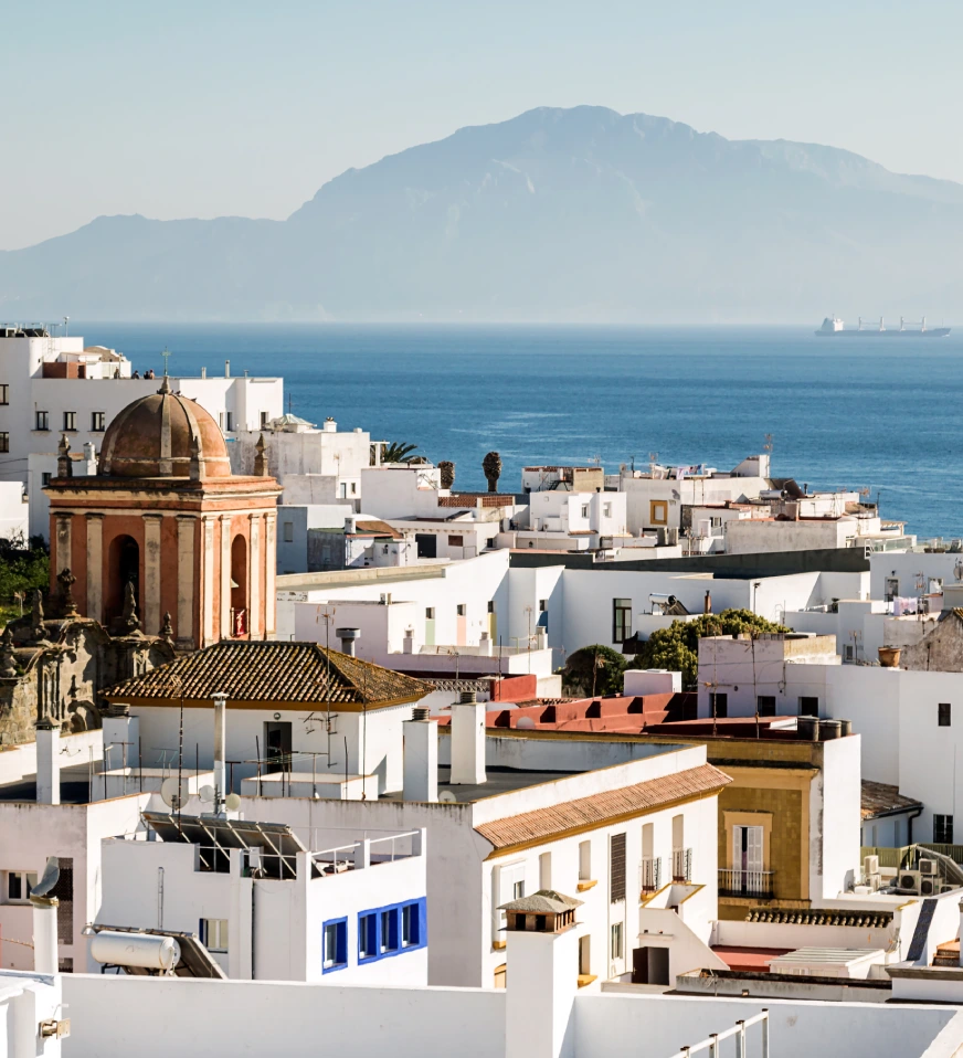 Vista de una ciudad costera con monta&ntilde;as al fondo y edificios blancos.
