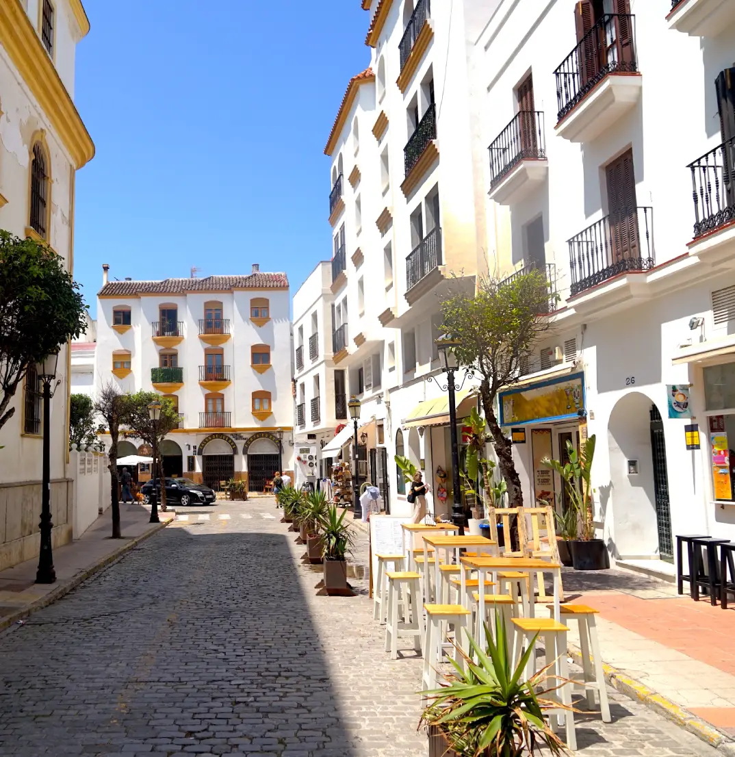 Calle adoquinada en ciudad mediterr&aacute;nea, con casas blancas y mesas al aire libre.