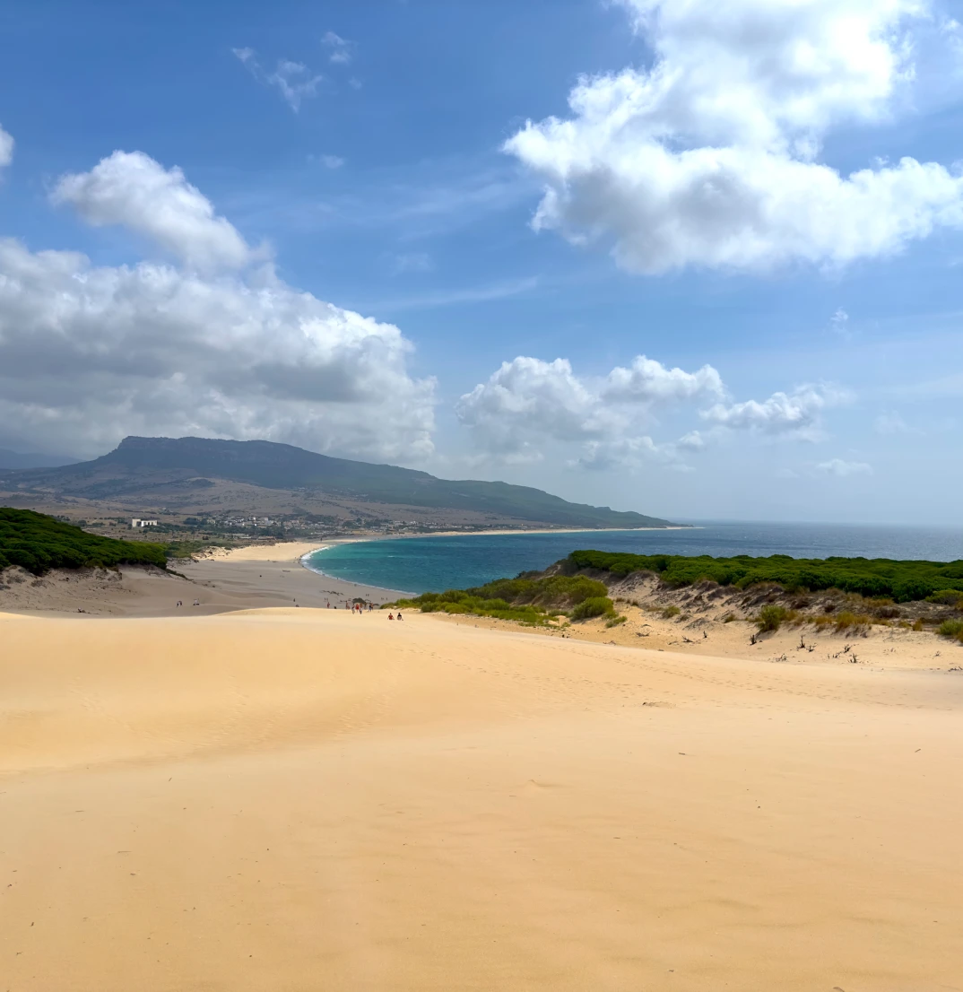 Playa con dunas de arena, mar azul y cielo parcialmente nublado.