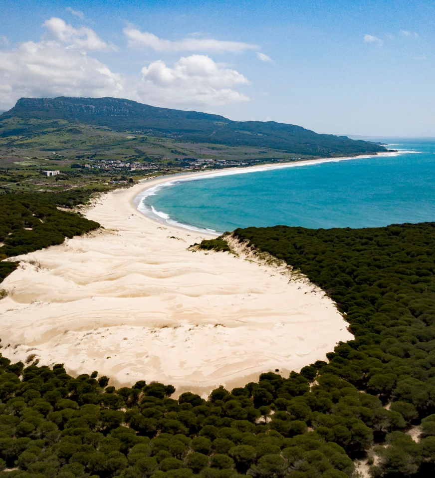 Vista a&eacute;rea de una playa con dunas, mar azul y monta&ntilde;as al fondo.
