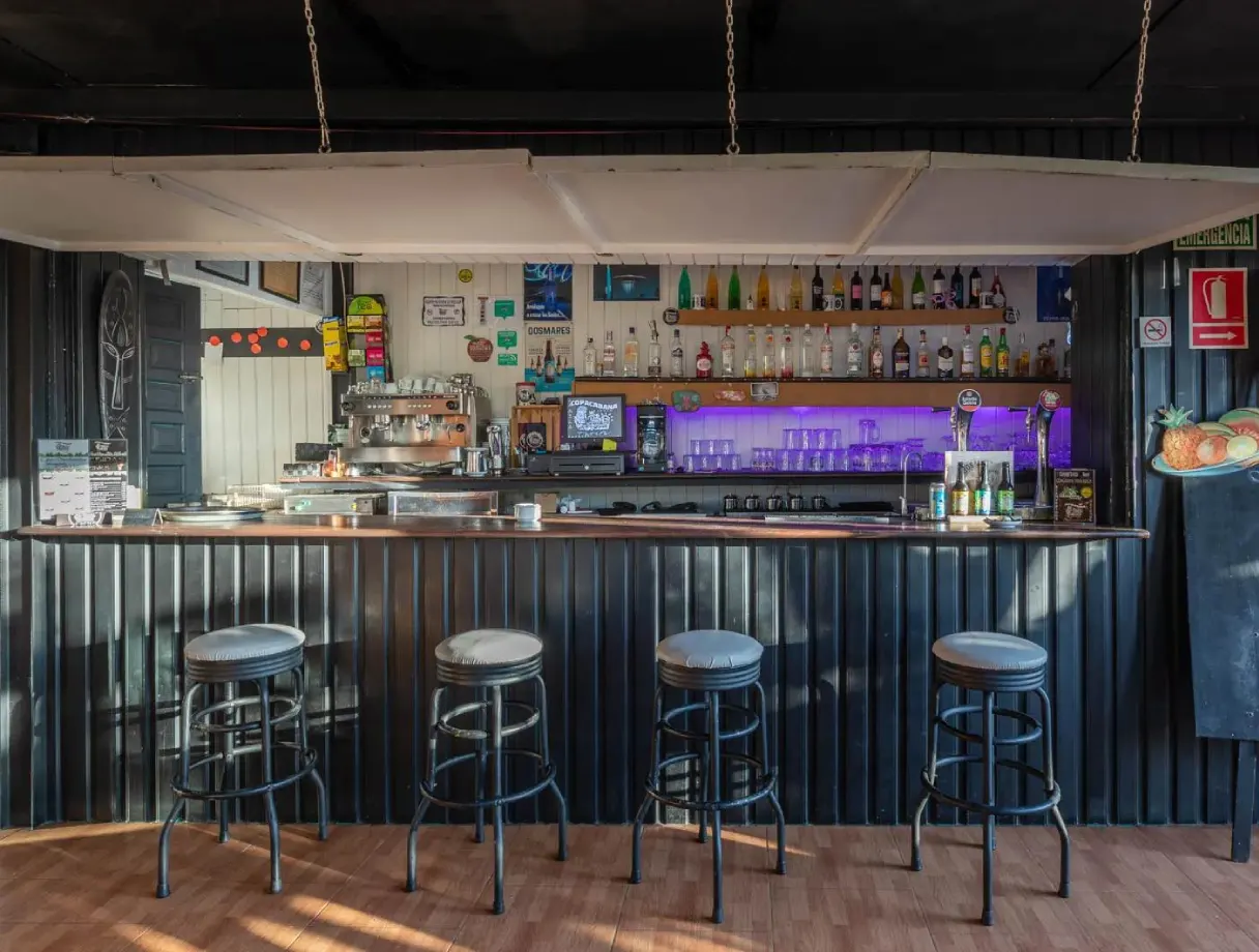 Bar con cuatro taburetes, botellas y luces moradas en la estanter&iacute;a trasera.