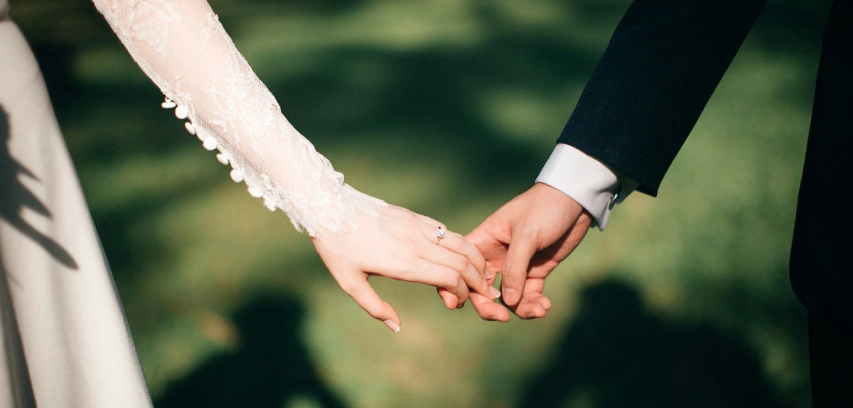 Manos de una pareja de novios entrelazadas sobre c&eacute;sped verde.