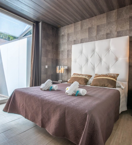 Cama con cabecero blanco, edred&oacute;n marr&oacute;n y toallas dobladas sobre la cama.