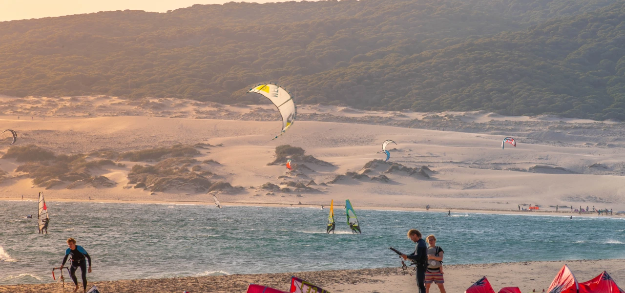 Personas practicando kitesurf en la playa con dunas y mar al fondo.