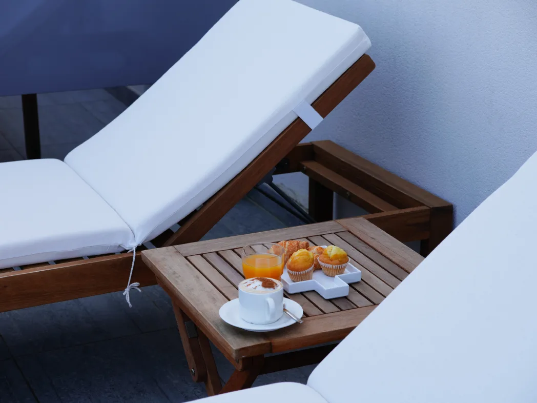 Mesa de madera con caf&eacute; y pasteles sobre tumbona junto a una pared blanca