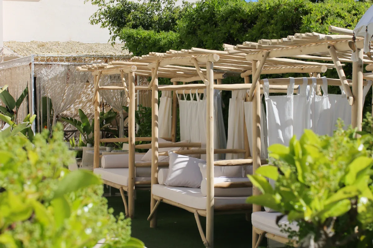 P&eacute;rgola de madera con camas y cortinas blancas, rodeada de vegetaci&oacute;n verde