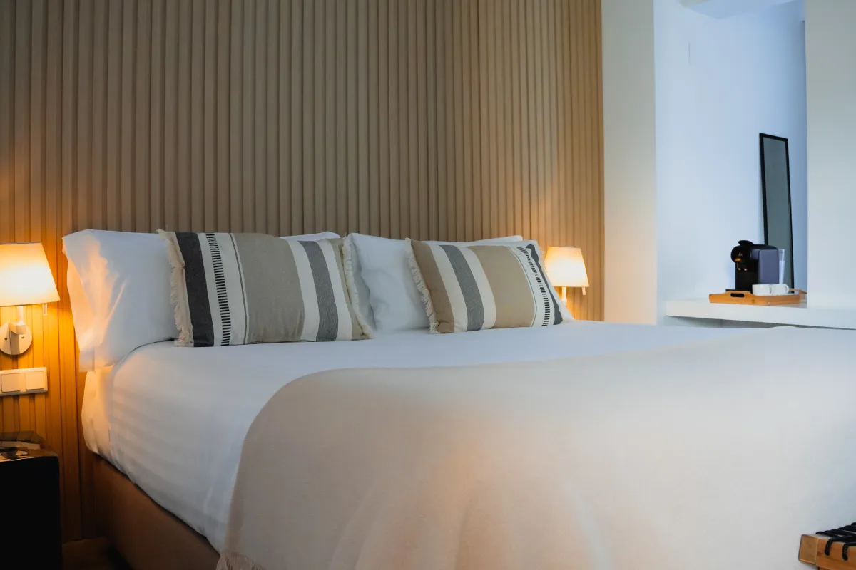 Cama matrimonial con almohadas y l&aacute;mparas, pared de madera y ventana azul