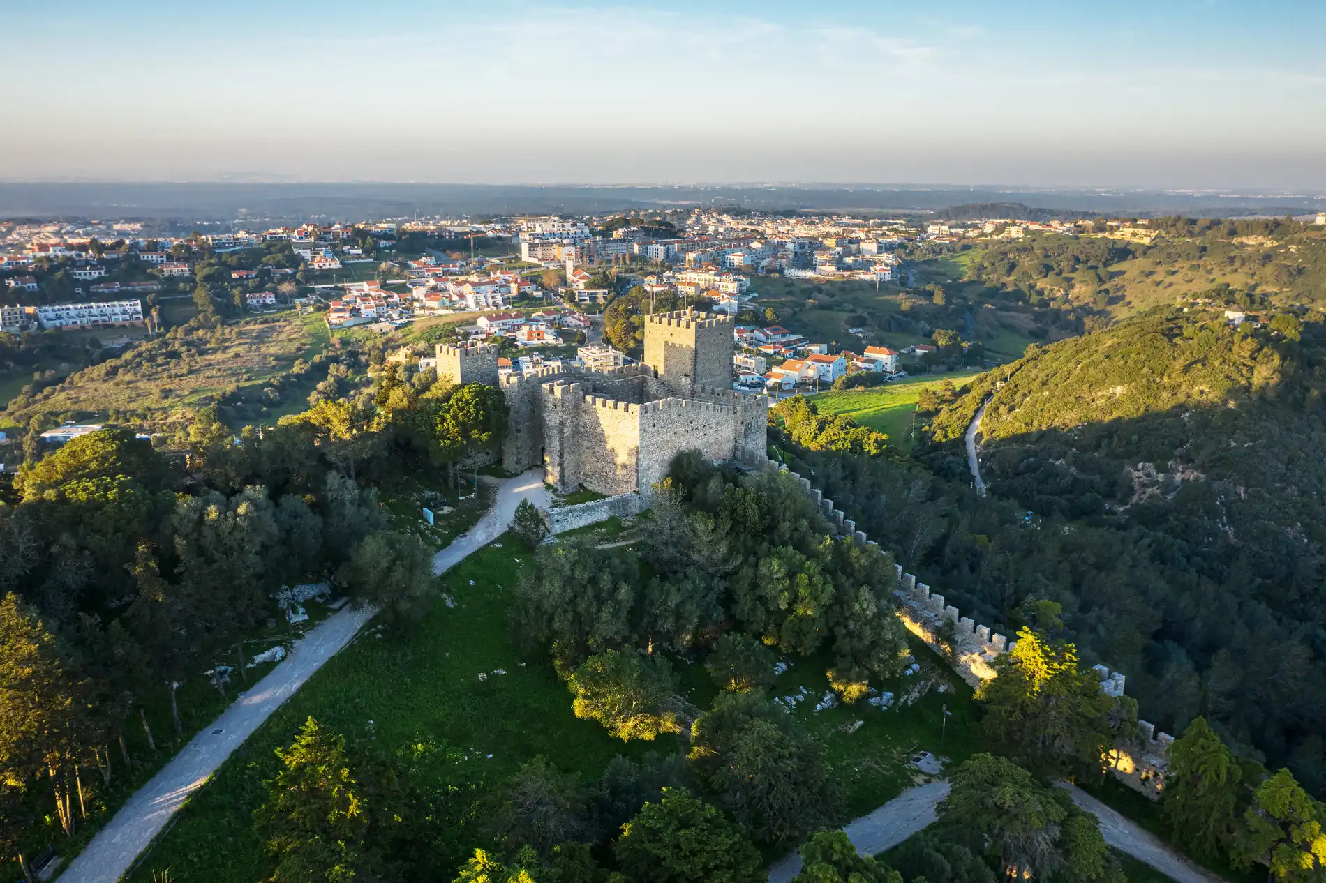 Castelo cercado por vegeta&ccedil;&atilde;o com cidade ao fundo, sob c&eacute;u claro.