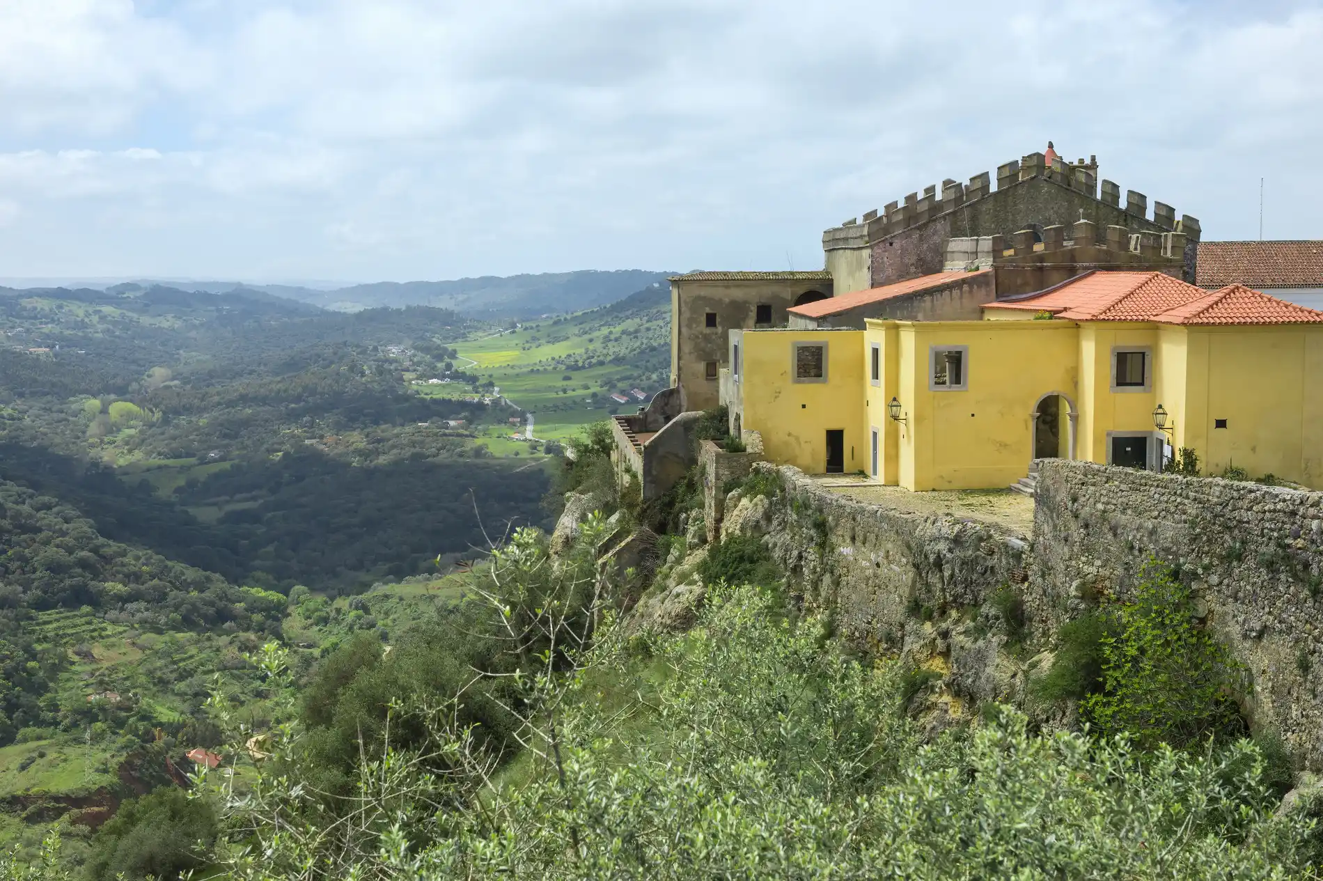 Casas amarelas em um penhasco, com vista para um vale verdejante e montanhas.