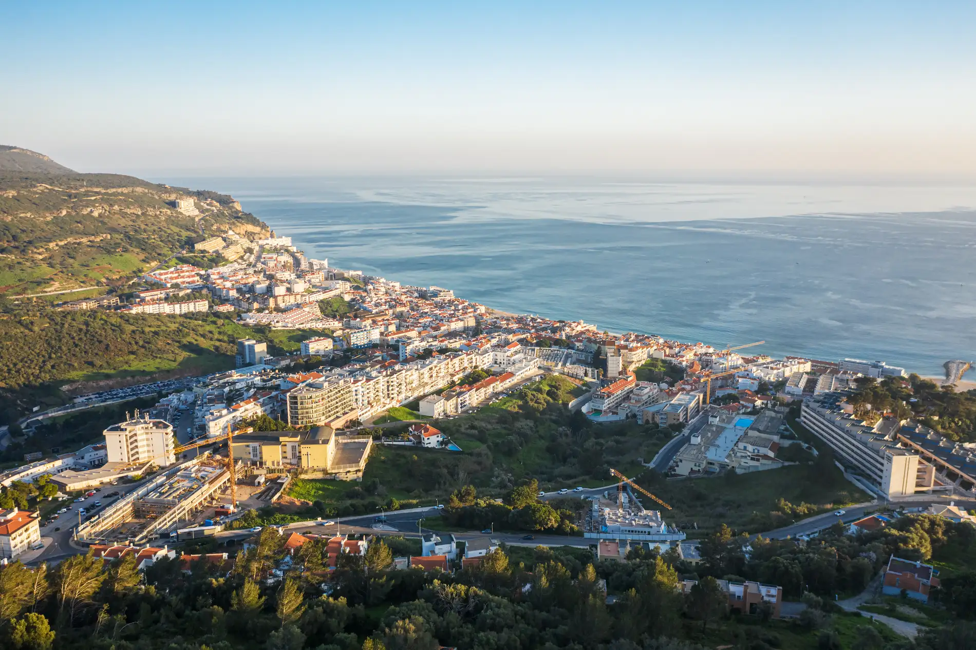 Vista a&eacute;rea de uma cidade costeira cercada por colinas e o mar ao fundo.