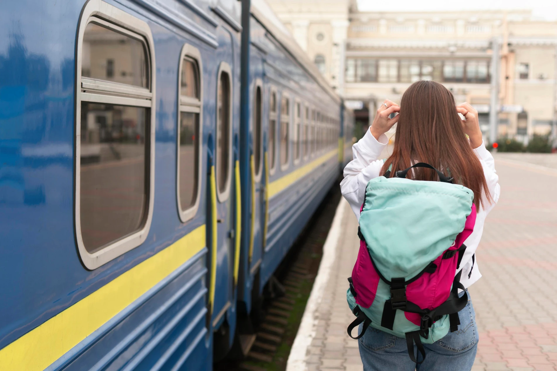 Mulher com mochila colorida ao lado de um trem azul na esta&ccedil;&atilde;o.