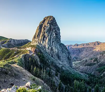 Tall rocky peak amidst green hills under a clear blue sky.