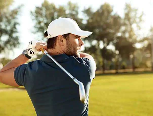 Golfer in a blue shirt and cap swinging a club on a sunny course.