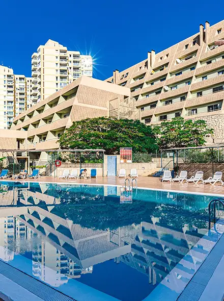 A sunny hotel pool with lounge chairs, surrounded by modern buildings.