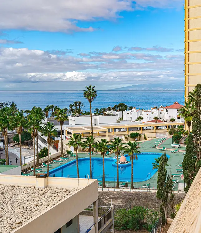 Coastal resort with a pool, palm trees, and ocean view under a blue sky.