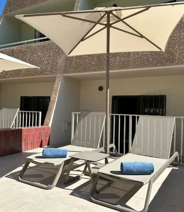 Two lounge chairs with blue towels under an umbrella on a sunlit patio.