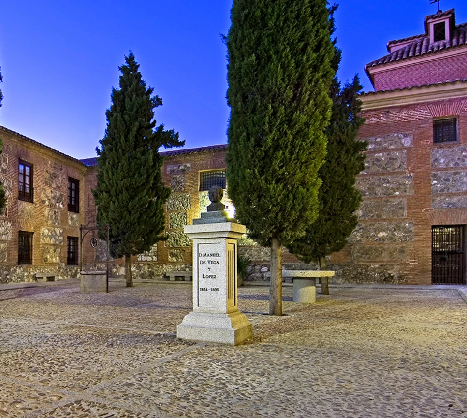 Plaza con monumento y &aacute;rboles al anochecer, edificio de ladrillo al fondo.