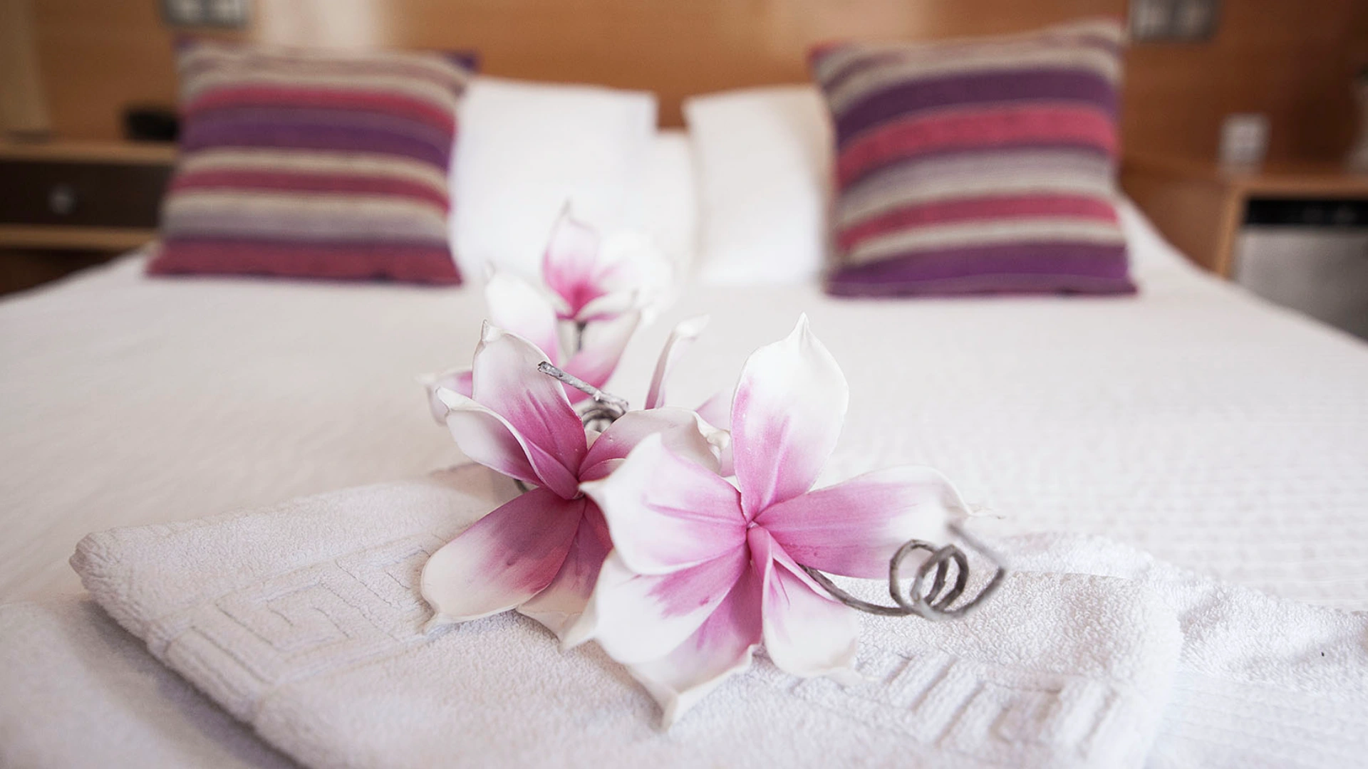 Toallas blancas con flores rosa sobre cama, cojines en el fondo.