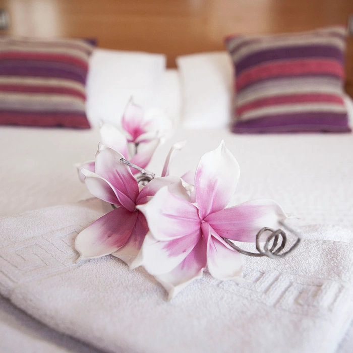 Flores rosadas sobre toallas blancas en una cama, con cojines a rayas atr&aacute;s.