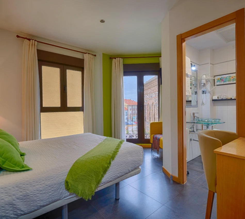 Habitaci&oacute;n con cama, escritorio y ba&ntilde;o, iluminada por luz natural.
