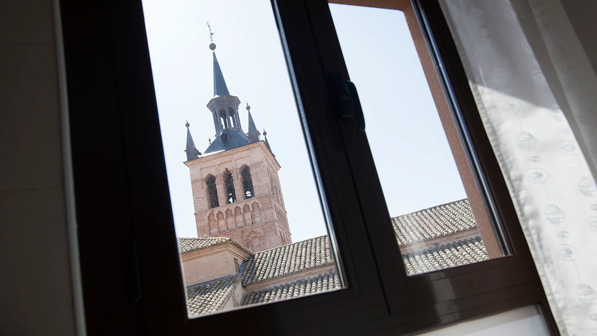 Torre de iglesia vista a trav&eacute;s de una ventana abierta con cortina blanca.