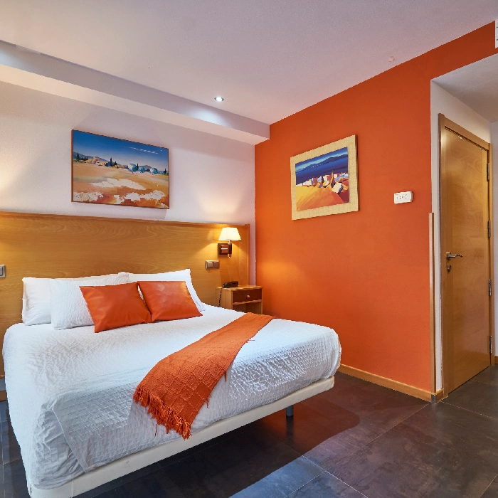 Habitaci&oacute;n moderna con paredes naranjas, cama blanca y cuadros coloridos.