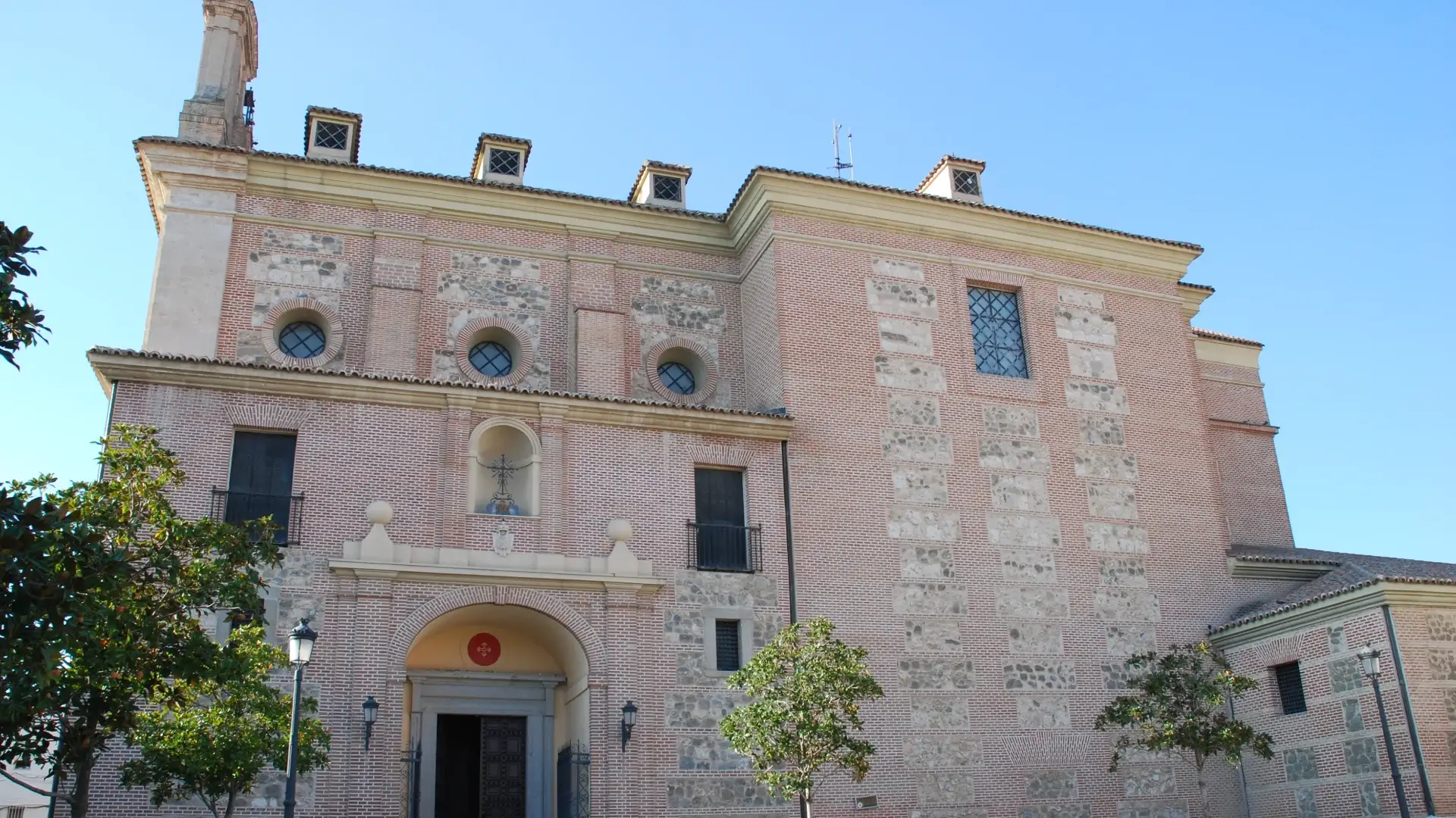 Edificio de ladrillo rojo con ventanas circulares y &aacute;rboles delante.